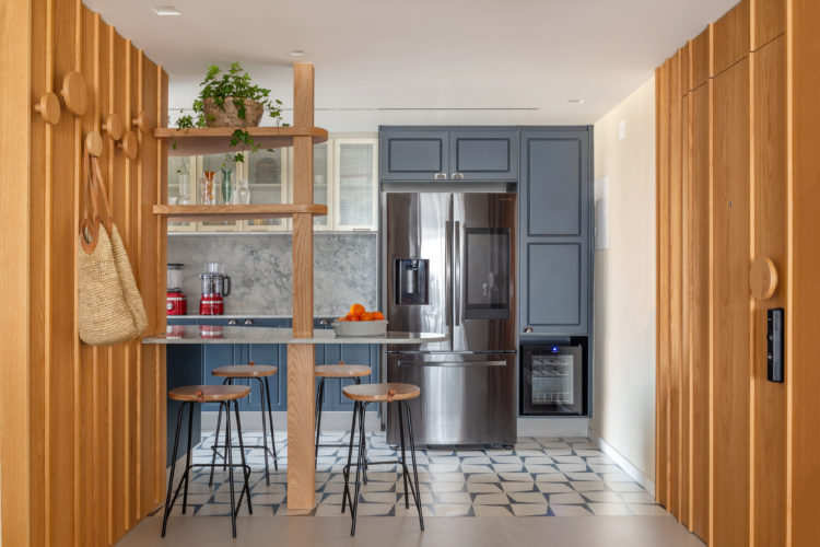Cozinha aberta para a sala, com piso em ladrilho hidráulico, armários na cor azul, grande geladeira em inox, e nas laterais, ripas em madeira cobrem a parede.
