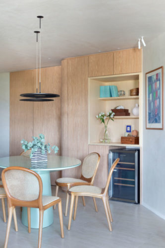 Living decorado e tons claros, parede forrada em madeira, mesa redonda na cor pistache, e cadeiras em palhinha. 