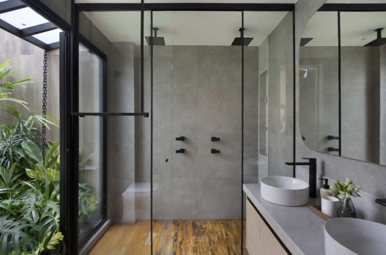 Banheiro revestido om porcelanato cinza, os dois chuveiros no box e as torneiras, na cor preta, ao fundo um jardim 
