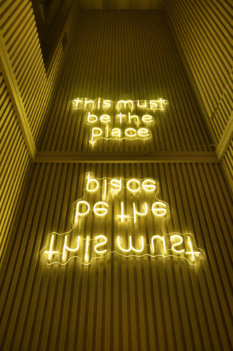 no hall do elevador, com paredes forradas de madeira ripada e teto espelhado para refletir a luminária com luz de neon que reproduz uma frase da música THIS MUST BE THE PLACE (do Talking Heads), fixada de cabeça para baixo.