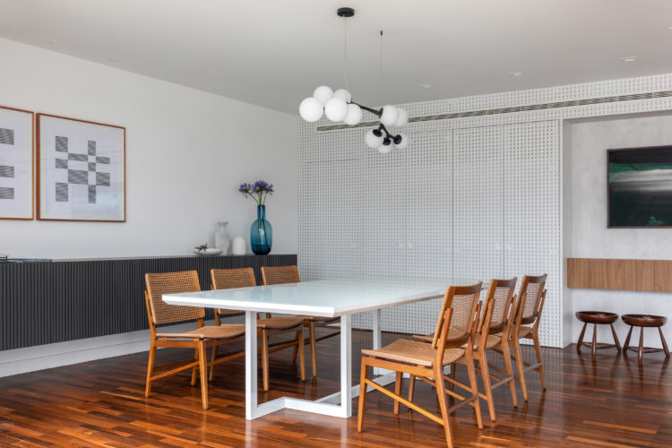 Ambiente de sala de jantar. Mesa branca e cadeiras em madeira, ao fundo, painel em madeira pintada de branco criando o efeito em muxarabi