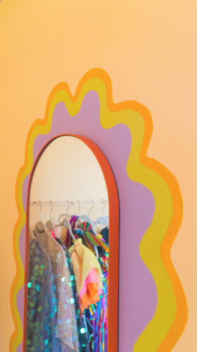 Dopamine Decor: Estilo de decoração que busca trazer, por meio do uso de muitas cores e padronagens divertidas, a sensação de felicidade para dentro de casa. Parede laranja com um espelho de corpo pendurado, e em volta dele uma pintura artística.