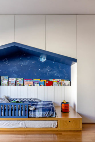 Quarto de criança, cama baixa e na parede, um nicho com desenho de teto da uma casinha. No fundo do nicho, pintura do céu com uma lua, e livros apoiados.