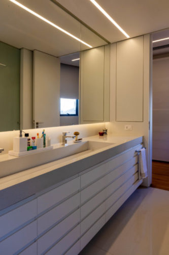 Banheiro com bancada e armários na cor branca, um grande espelho em toda a extensão da bancada, 