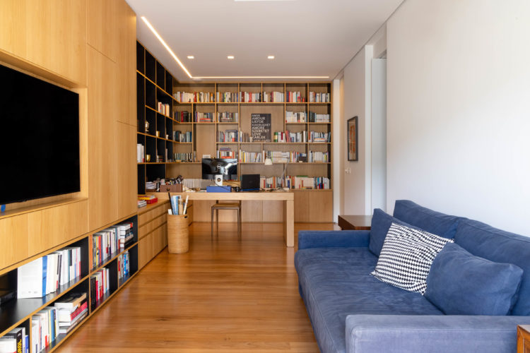 Home office, piso em madeira, uma estante grande repleta de nichos com livros, bancada em madeira, um painel na parede toda, também em madeira, abriga uma tv embutida