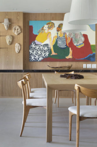 Sala de jantar. Ambiente revestido em madeira, mesa também em madeira, uma tela colorida ao fundo, e dois pendentes brancos em cima da mesa; 