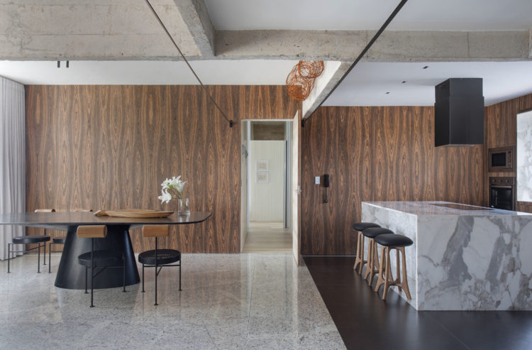 Cozinha e sala totalmente integradas. Parede de fundo forrada em madeira escura, pau ferro, vigas em concreto aparente.