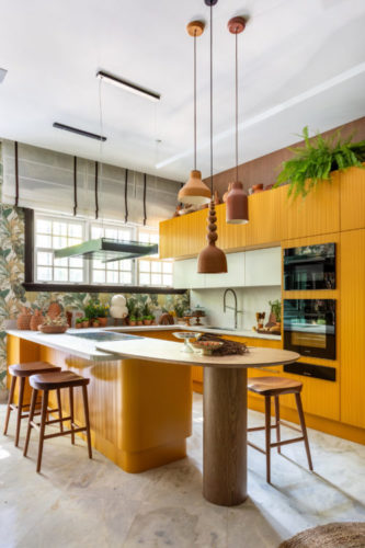 Cozinha na mostra CasaCor Rio 2023. Papel de parede estampado e armários amarelos. Bancada alta siando da ilha com cooktop