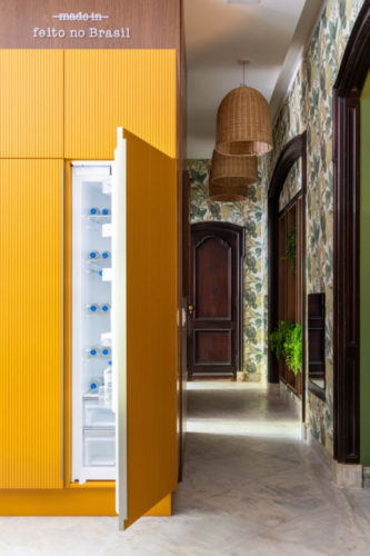 Feito no Brasil; frase em cima do armário amarelo que oculta a geladeira. Cozinha na mostra CasaCor Rio 2023. Papel de parede estampado e armários amarelos.