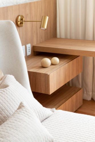 Detalhe de marcenaria, a mesa de cabeceira acoplada ao painel de madeira atras da cama, e saindo dele, uma bancada também em madeira.