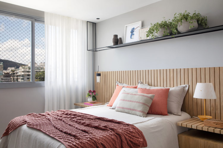 Mix de materiais marca o décor deste apartamento de 110m2, em Niterói. No quarto de casal, cabeceira da cama e mesas laterais em madeira ripada, cortina em linho branco, e uma prateleira em cima da cama com serralhieria na cor preta.
