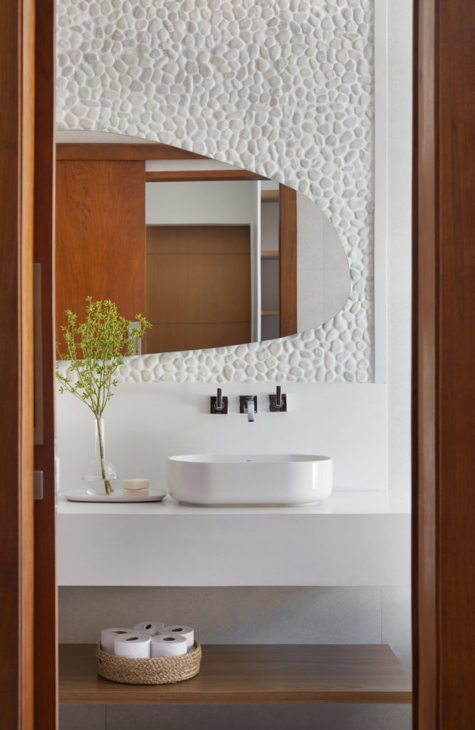 Lavabo com revestimento em pedras,seixos rolados, na parede da cuba, espelho em formato organico. Bancada branca.