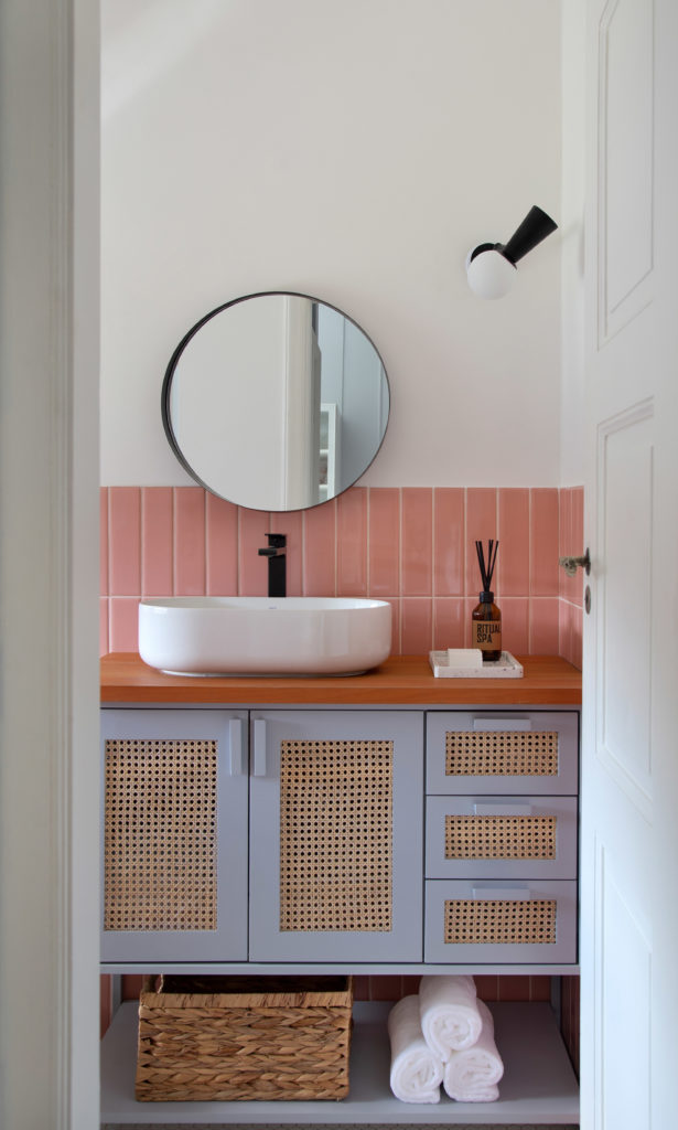 Banheiro com meia parede em revestimento na cor rosa, e pintado de branco na outra parte. Armarios azuis claros com detalhes em palhinha