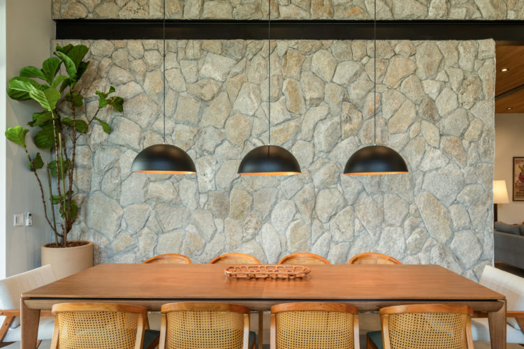Ambiente de sala de jantar, com mesa em madeira, cadeira em plhinha e madeira, três pendentes em formato de meio circulo, e aon fundo, parede revestida em pedra.