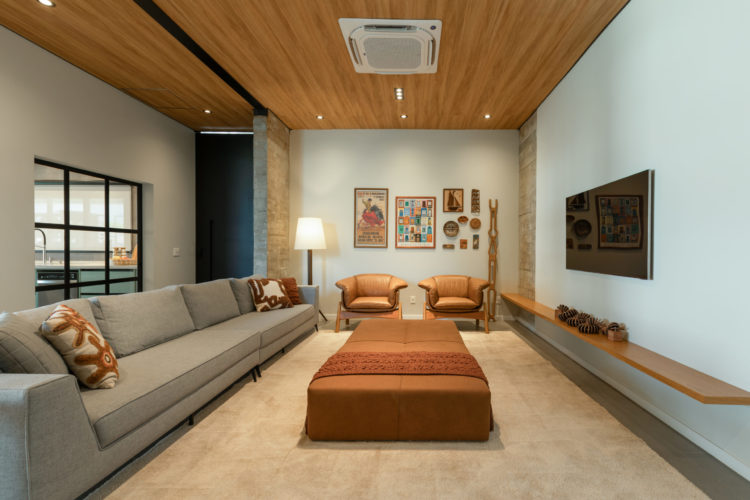 Sala de Tv em uma casa de campo no interior de São Paulo. Sala com layout retangular, um grande sofá cinza claro em frente a tv que está na parere, No centro, um grande puff na cor marrom.