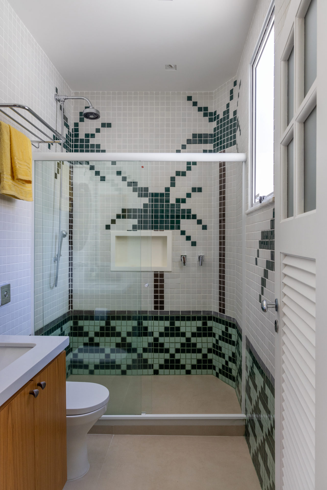 Banheiro revestido com pastilhas brancas e tons de verde, que formam desenhos de palmeiras