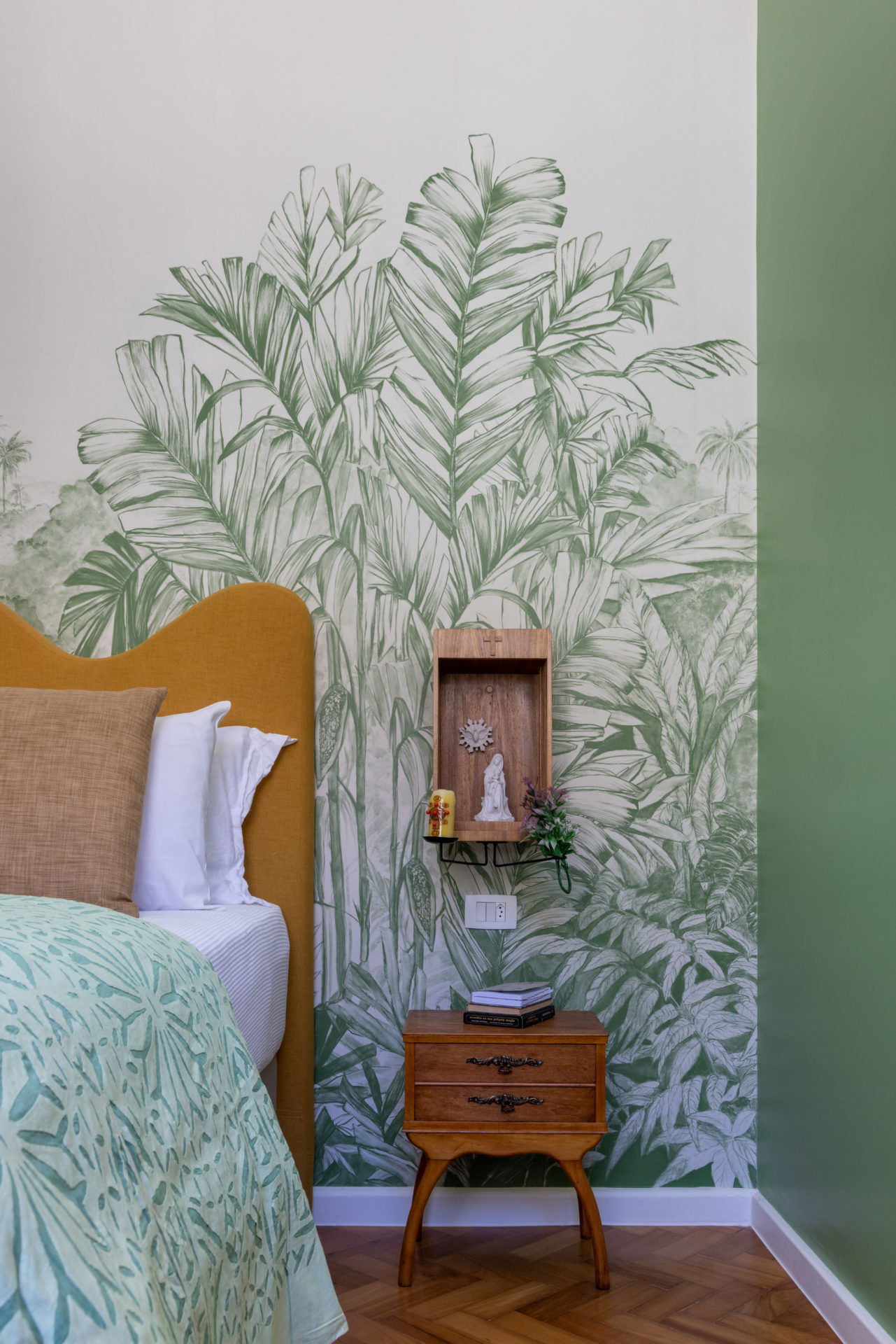 Cabeceira amarela, e na parede atras da cama, papel de parede branco e verde, com folhagens. Ao lado da cama, um oratorio em madeira