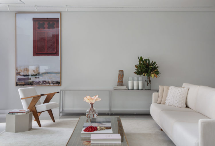 Living decorado em tons claros, piso, parede, sofá e poltrona na cor branca, com destaque para uma fotografia de uma janela vermelha