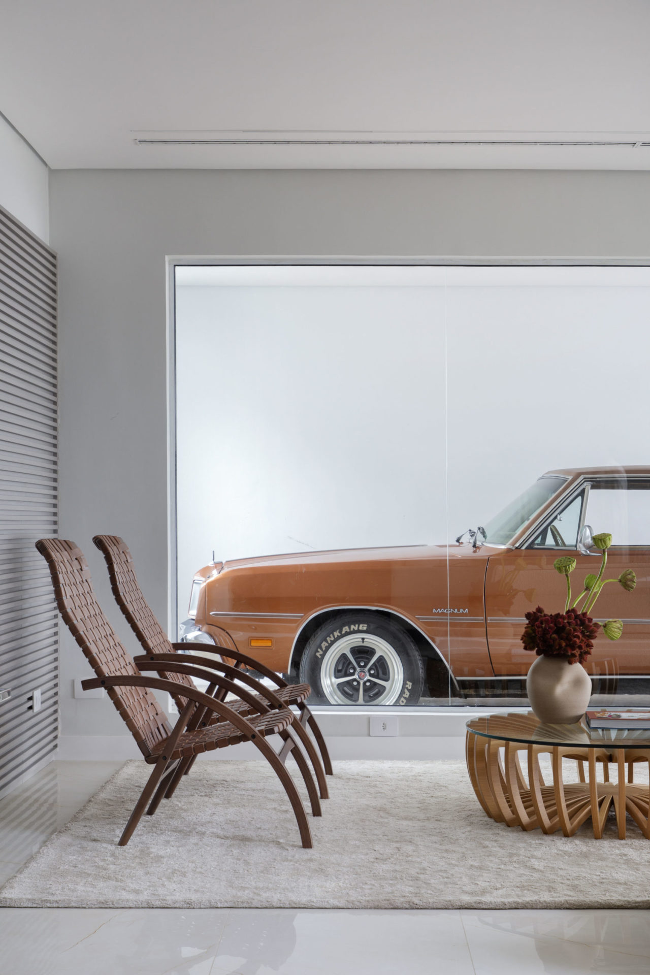 Peças de mobiliario com design assinado, em uma sala com um grande painel em vidro, de onde se vê a garagem, com um carro antigo estacionado