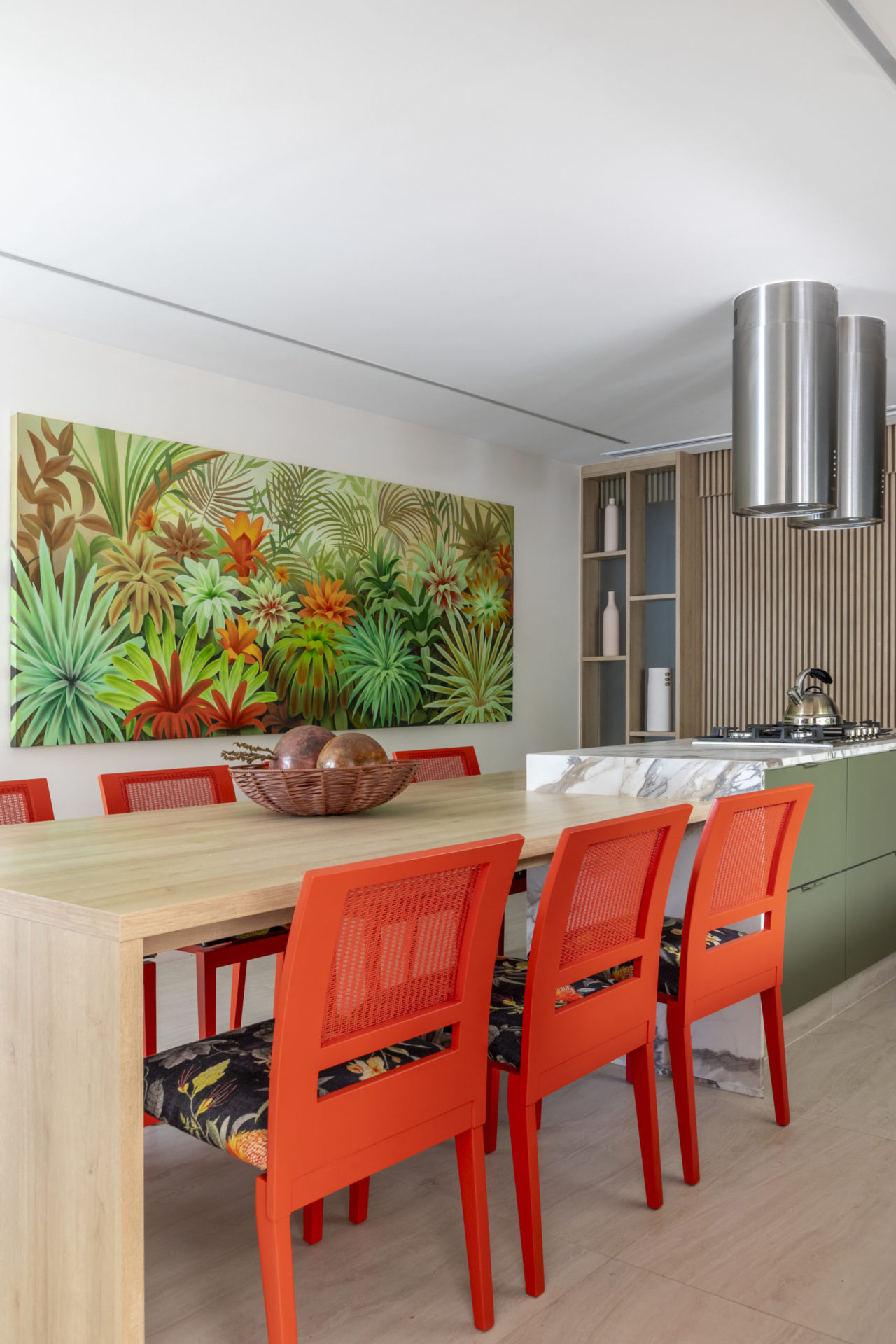 Cozinha com ilha central, duas coifas redondas, uma mesa e cadeiras na cor laranja, acoplada a ilha, e ao fundo, um quadro repelta de bromélias decorão a cozinha
