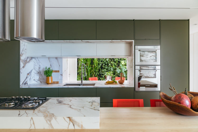 Cozinha com armarios na cor verde, com um grande nicho revestido em mármore, que é aberto para o ambiente da lavanderia, que tem um cantinho com parede verde