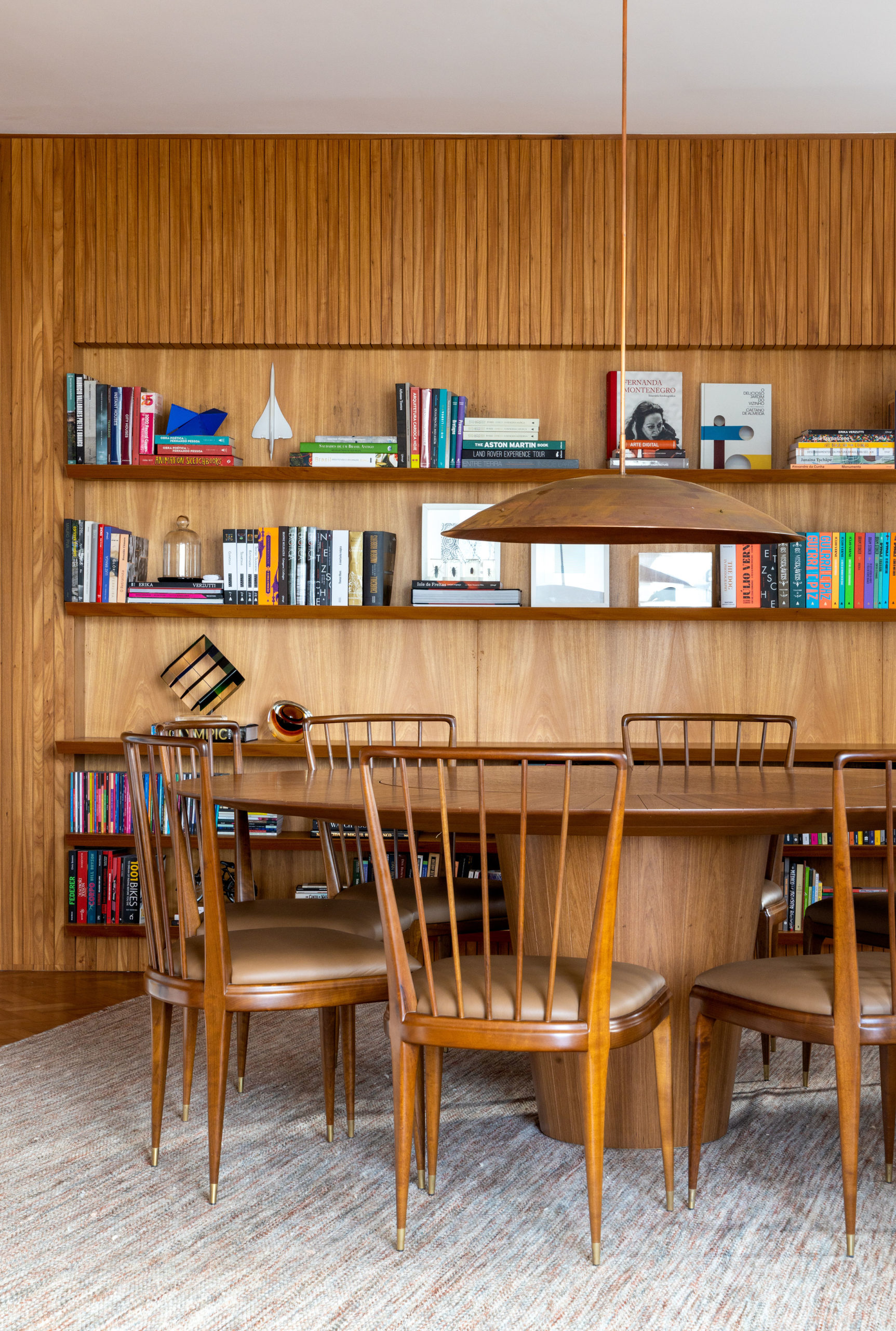 Ambiente da sala de jantar, com a parede de fundo revestida em madeira e prateliras para livros e uma delas servindo como aparador. Mesa em madeira, mesa de jantar Disco (de Pedro Useche) e as cadeiras de jantar (de Giuseppe Scapinelli).
