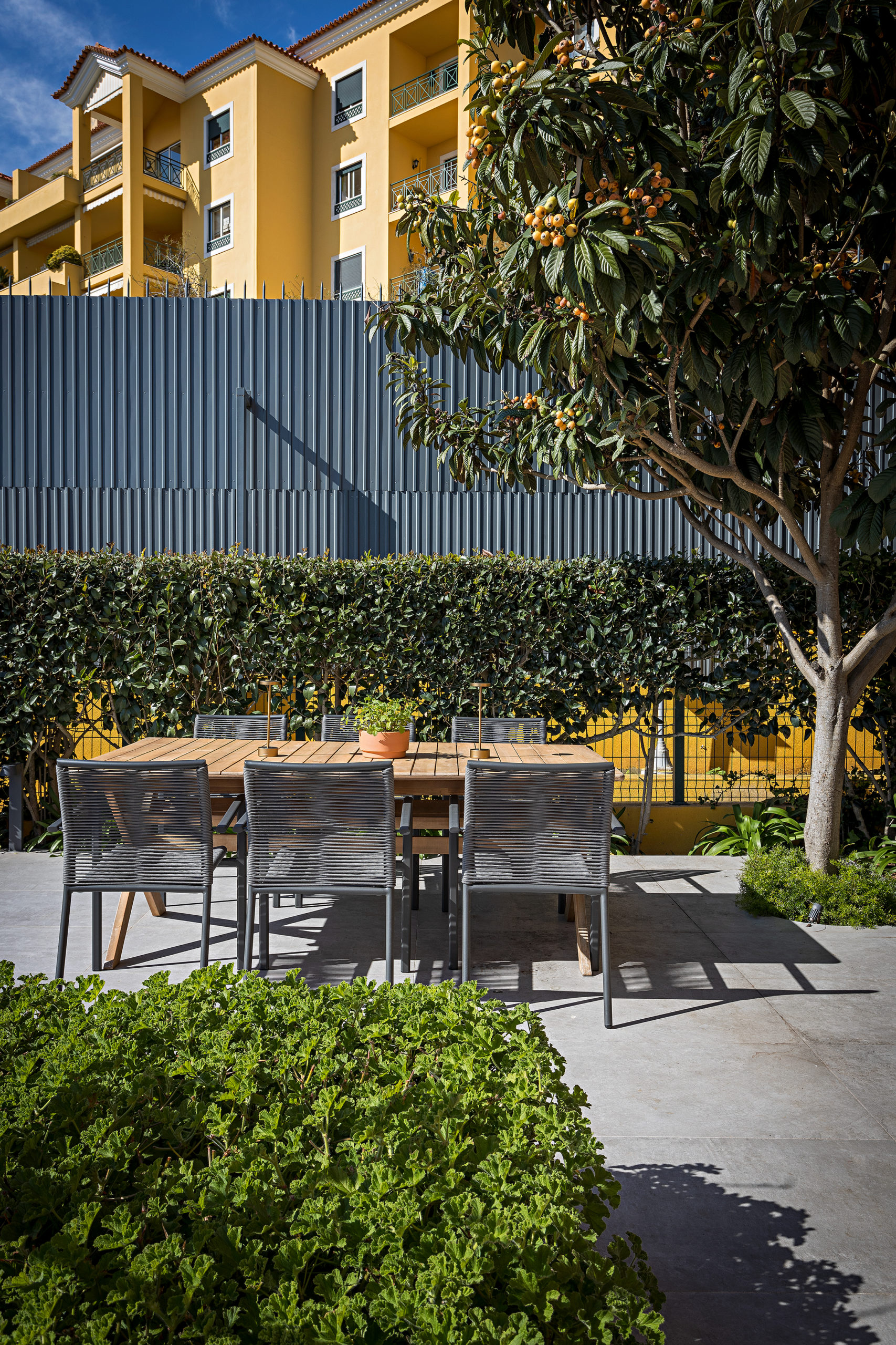 Área externa em uma casa em Estoril, Portugal, com uma mesa de jantar em madeira, e uma árvore e cercas vivas em volta.