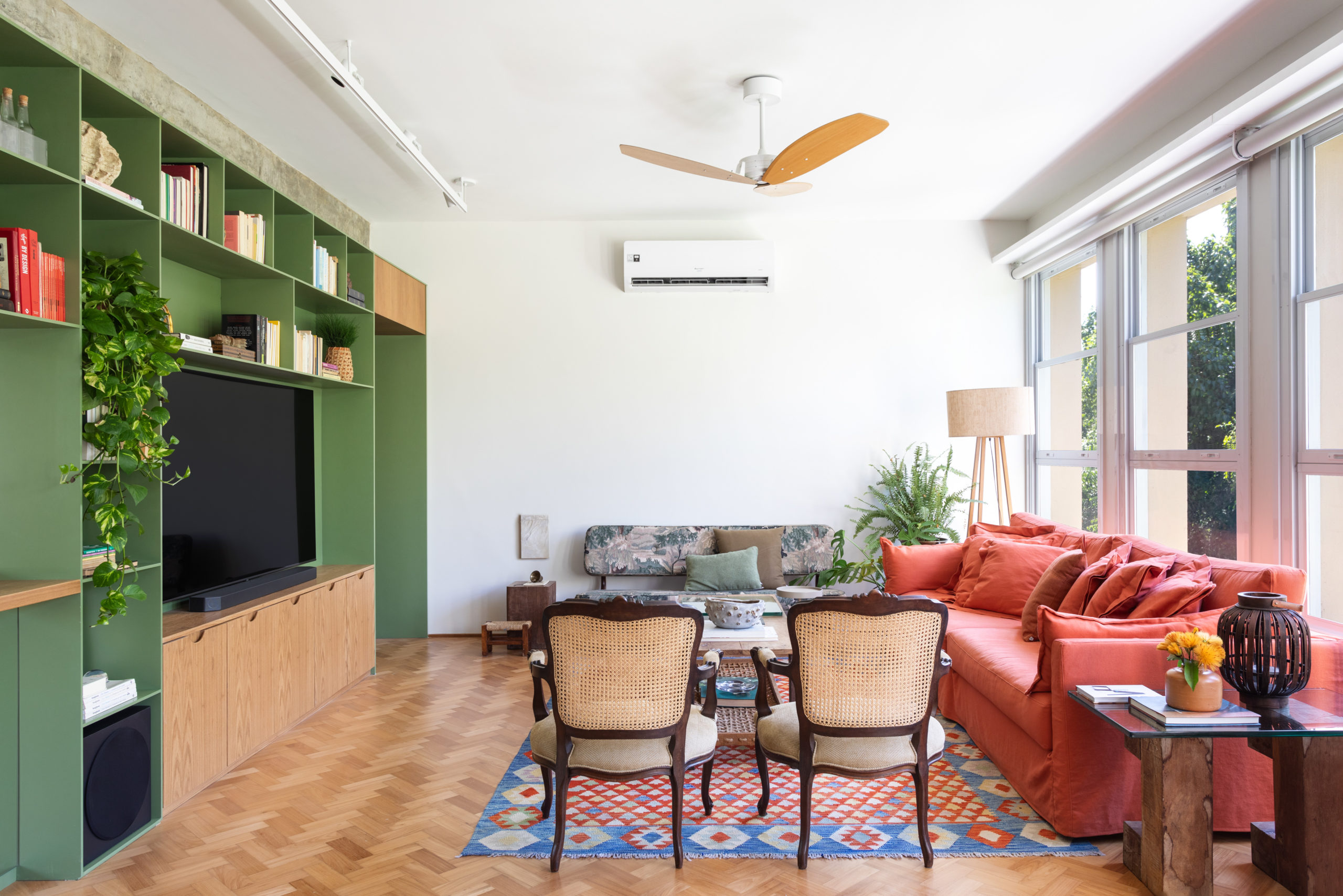 Sala ampla e clara, em um apartamento com janelas em todo a comprimento. Estante verde na parede em frente as janelas.