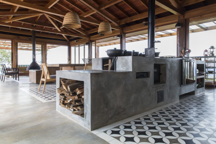 Na sala principal, um fogão à lenha aquece refeições em panelas de barro, destacando-se uma mesa de madeira robusta no espaço de jantar, cercada por ladrilho hidráulico que compõe o tapete desenhada pela Sabella Arquitetura