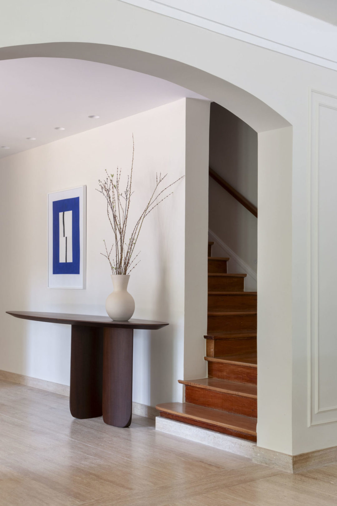 Hall de entrada de uma casa com arquitetura clássica, em Botafogo. Passagem da sala para o hall com um arco na parede, piso em mármore e escada para o segundo andar em madeira.