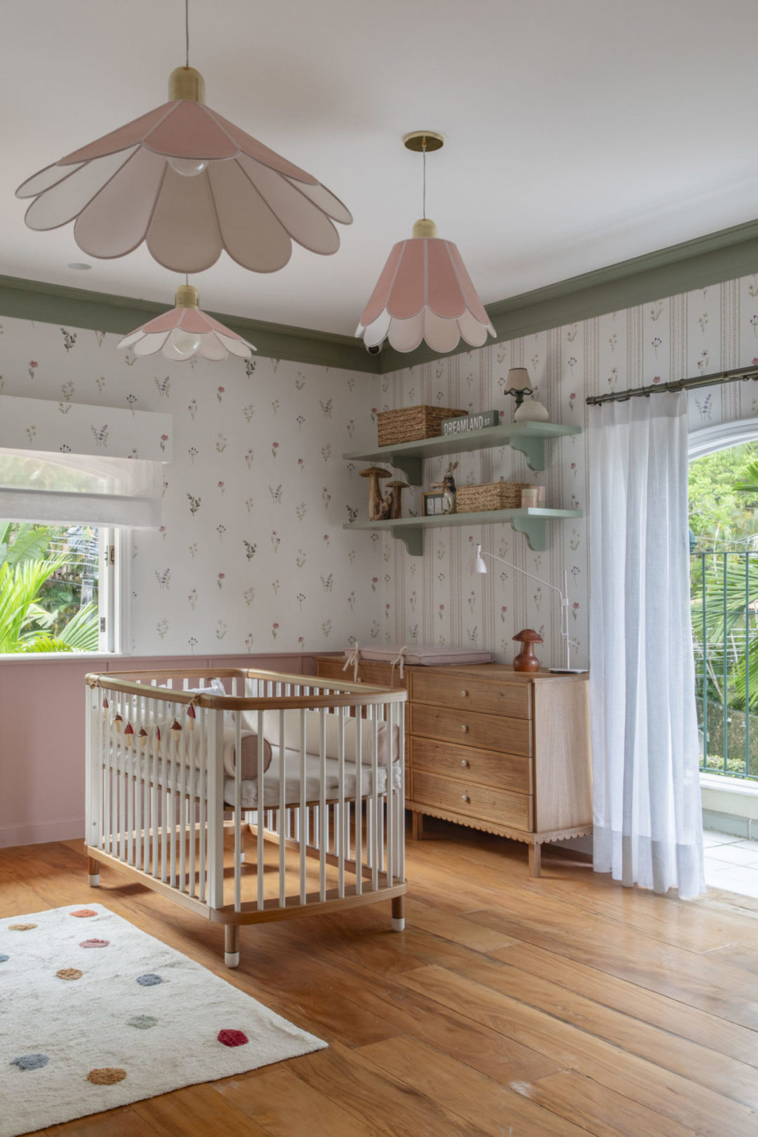 Quarto de bebê menina; piso em madeira, paredes com papel de parede floral e três laminárias pendentes em forma de flor.