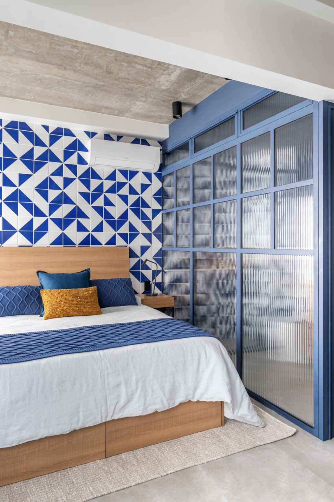 Parede atrás da cama, revestida de azulejo estampado de azul e branco. Ao lado, uma esquadria azul bebe com vidros canelados, divide o quarto do banheiro.