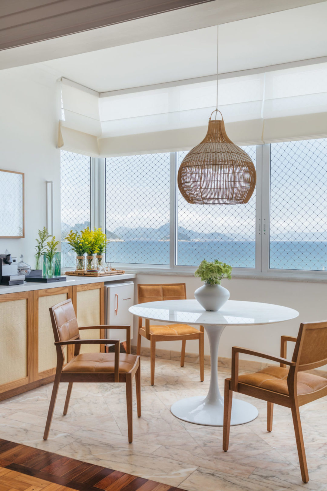 Ambiente em frente ao mar, com mesa redonda Sarinem, cadeiras em madeira, pendente em palha, e ao lado, aparador com mini geladeira embutida