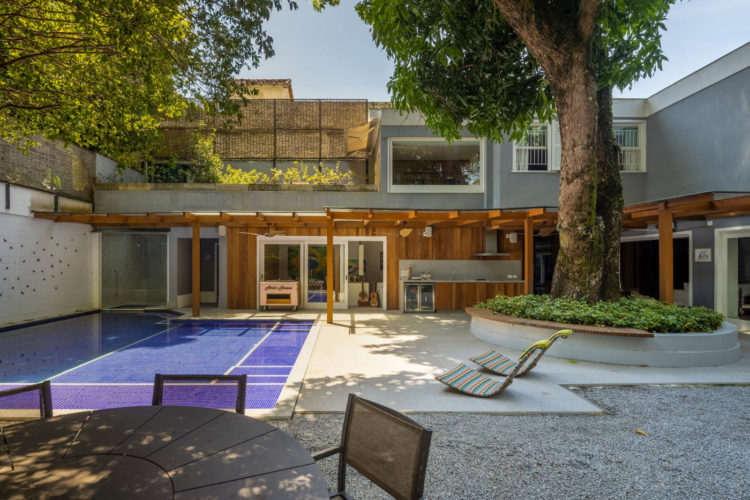 Área externa, em casa no Rio, com piscina, a´área gourmet e uma grande árvore com um banco de madeira ao redor.