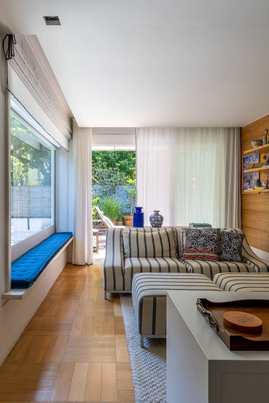 Saleta de um quarto de casal, com sofás em tecido listrado de branco e azul, ao fundo, um terraço que atende ao quarto.