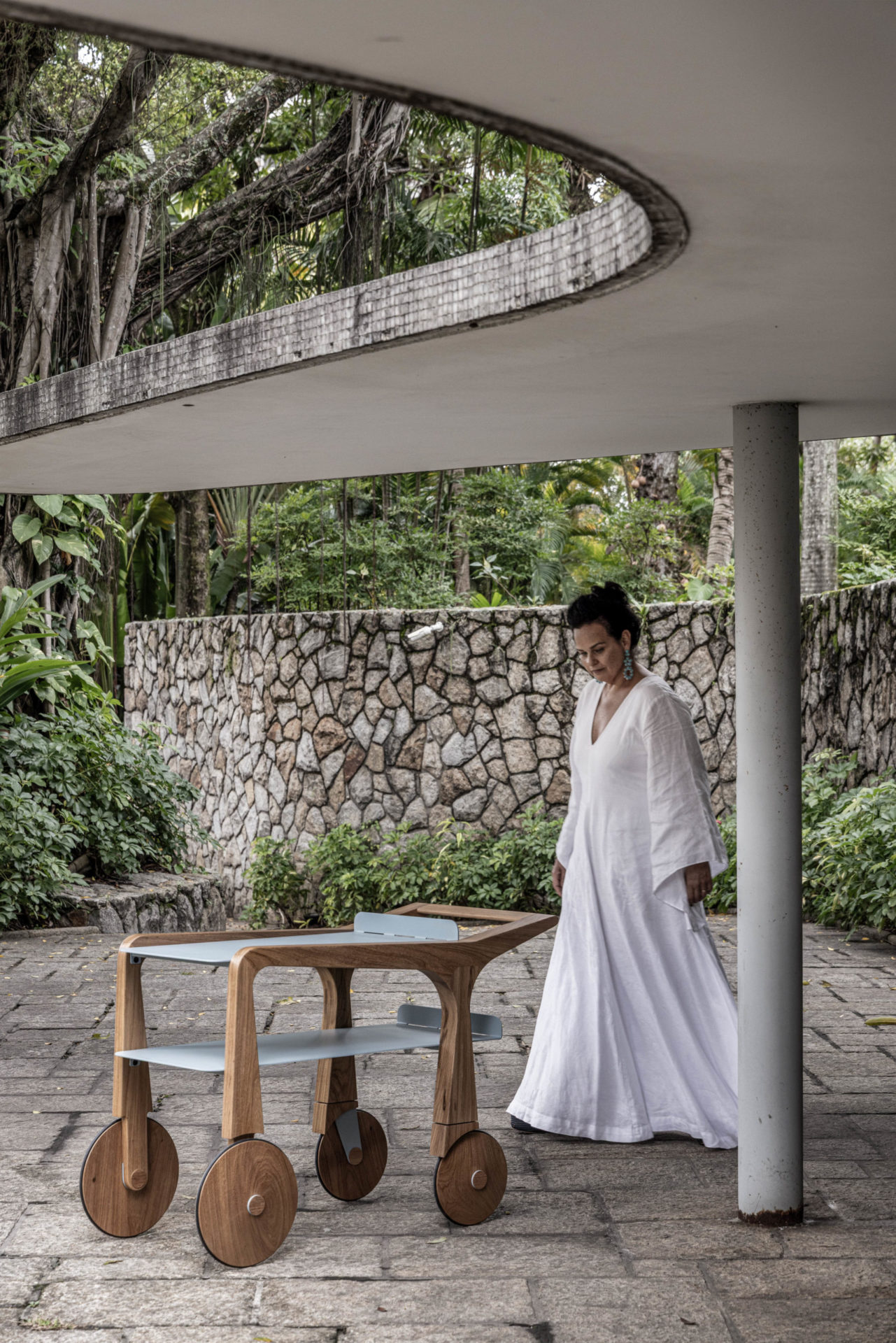 na foto mulher com roupa branca em área externa de uma casa com muito verde e um móvel de madeira
