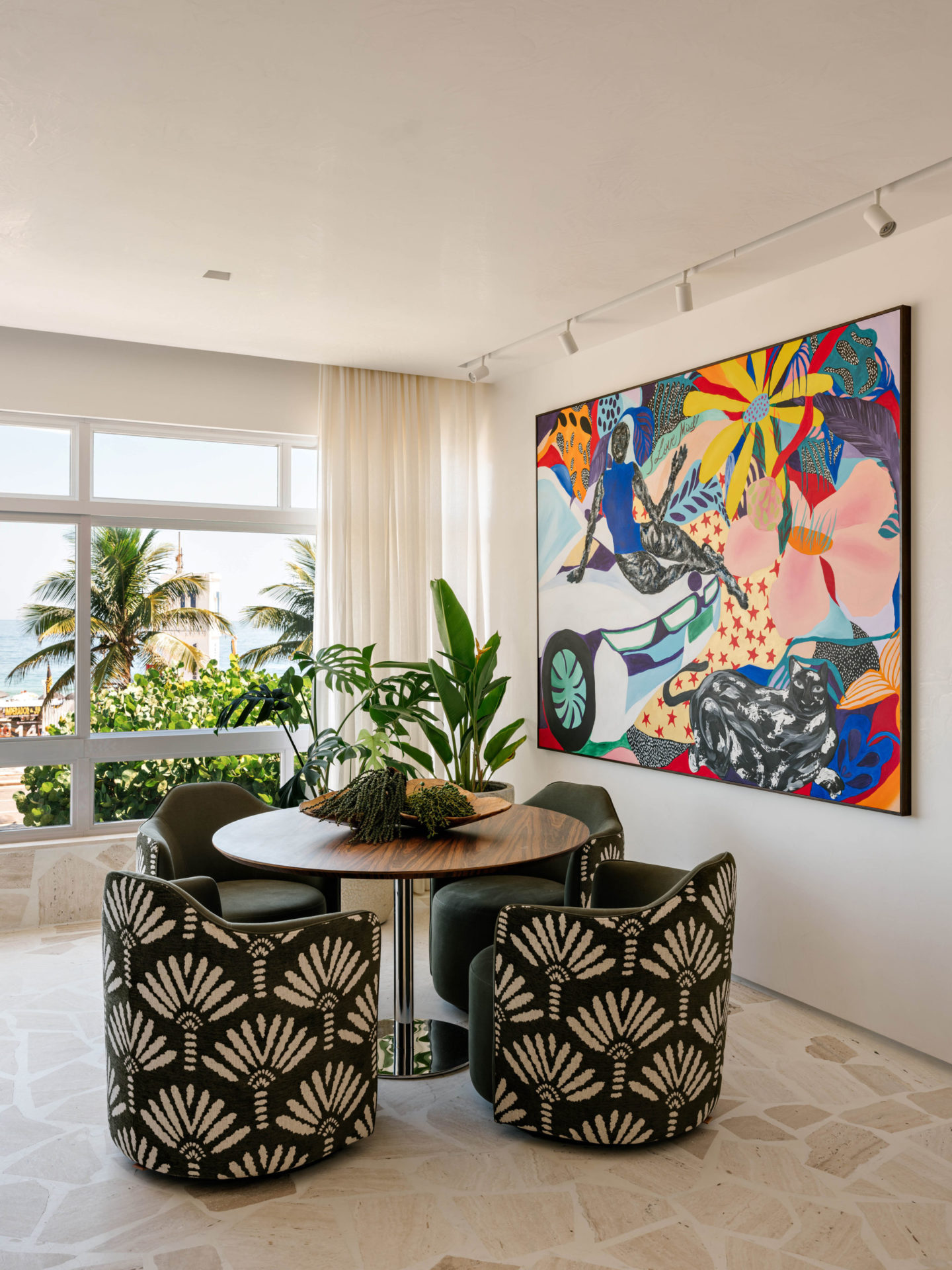 na foto uma sala com janelas grandes, uma mesa pequena com cadeiras e um quadro de muitas flores, folhas e cores