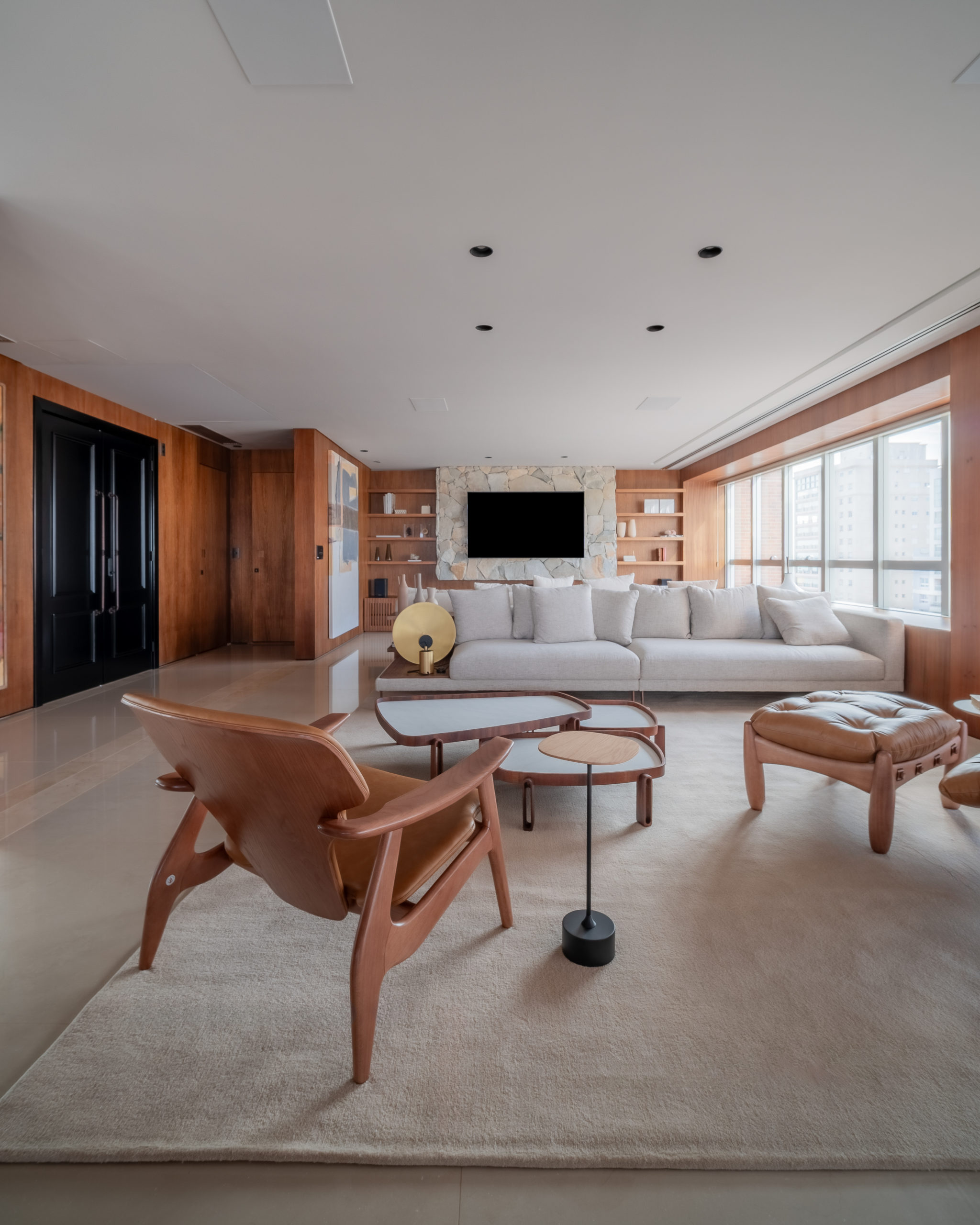 sala de estar com porta preta, paredes em madeira, sofá branco, poltrona de couro e tv na parede