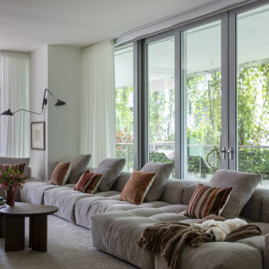 sala com sofá ilha em tom claro, almofadas, mesa de centro, luminária no canto da parede e varanda