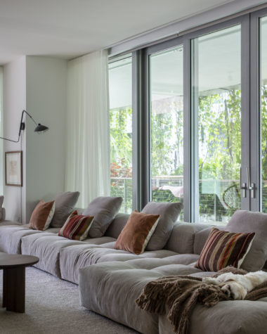 sala com sofá ilha em tom claro, almofadas, mesa de centro, luminária no canto da parede e varanda