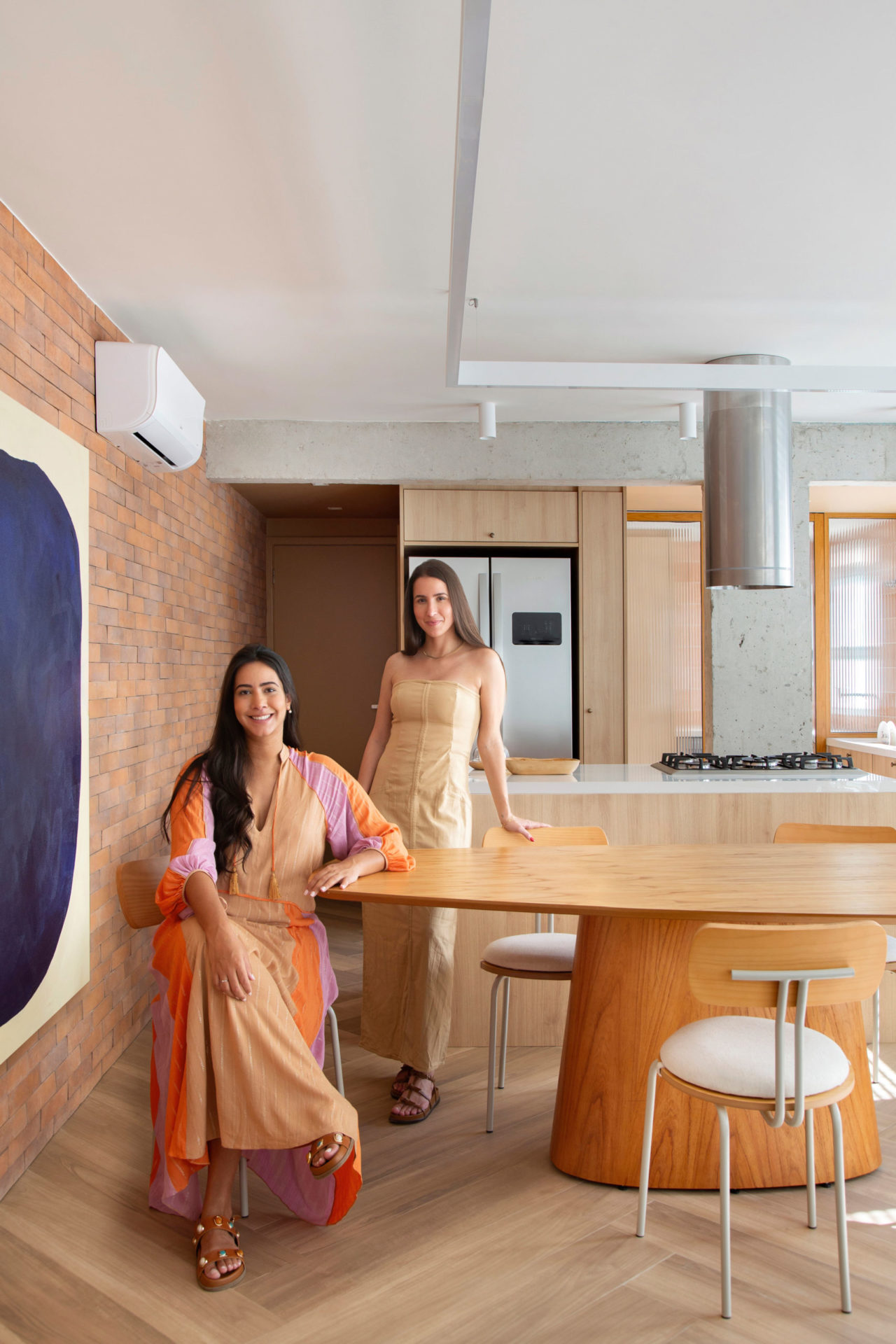 Uma mulher branca e de cabelos castanhos longos sentada e uma mulher branca em pé encostada na mesa de jantar