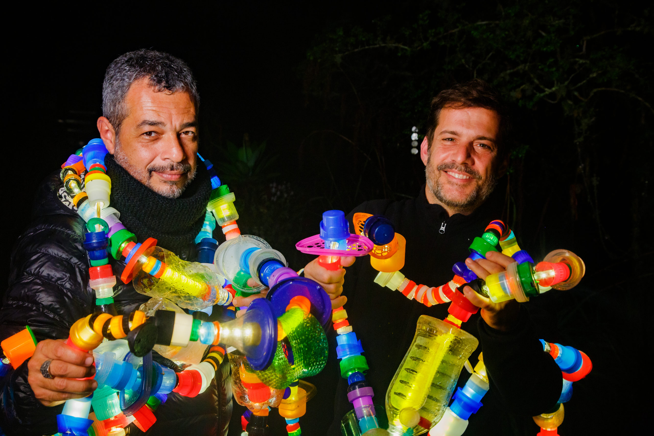 dois homens brancos em um fundo preto com luminárias coloridas feitas de reciclagem