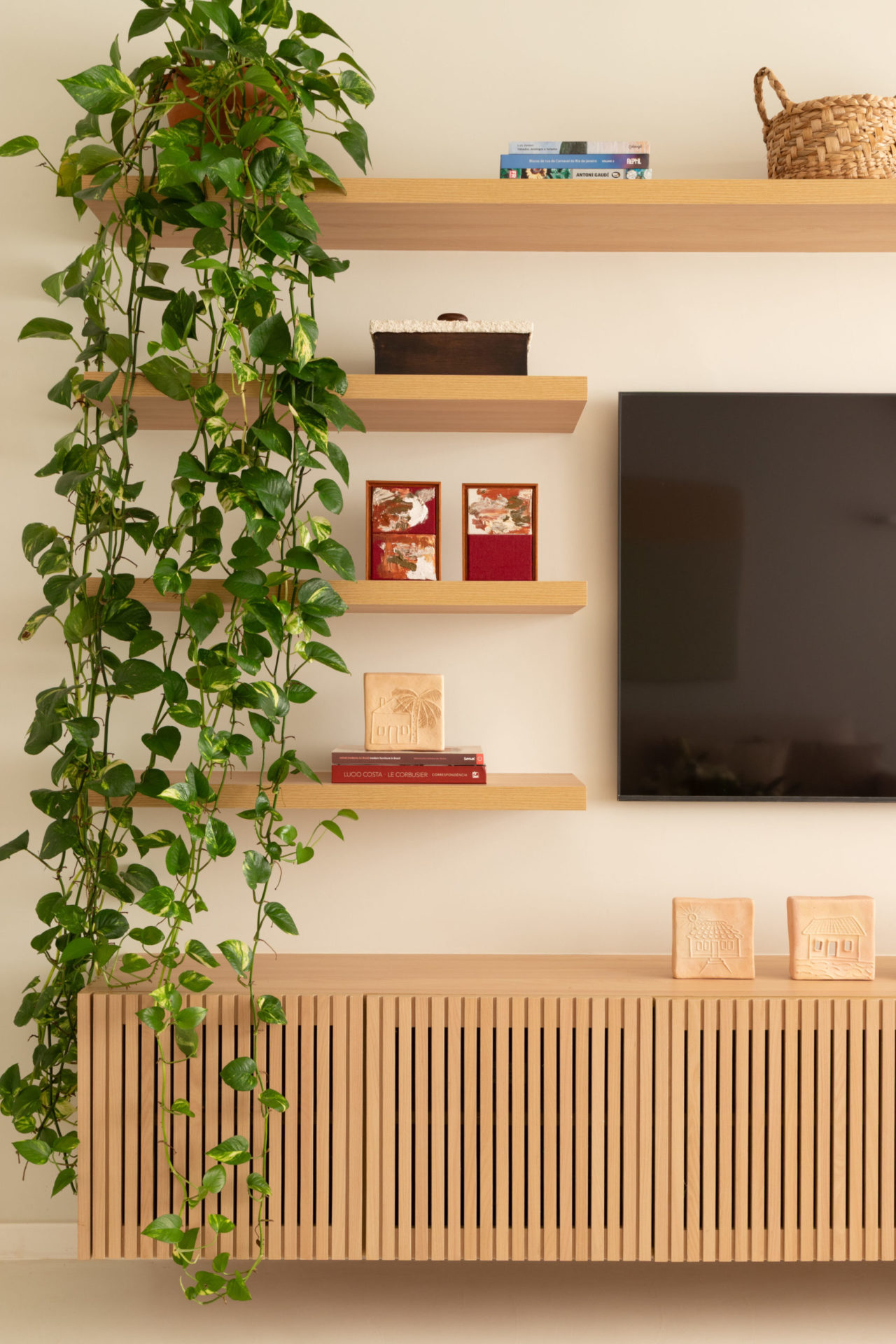 na foto uma sala com tv na parede, plantas e prateleiras em tons claros