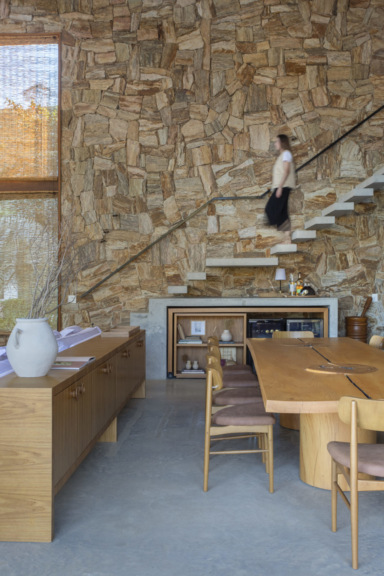 na foto é possível ver interior de casa com pedra natural nas paredes e madeira cumaru no forro, uma escada e mesa de madeira
