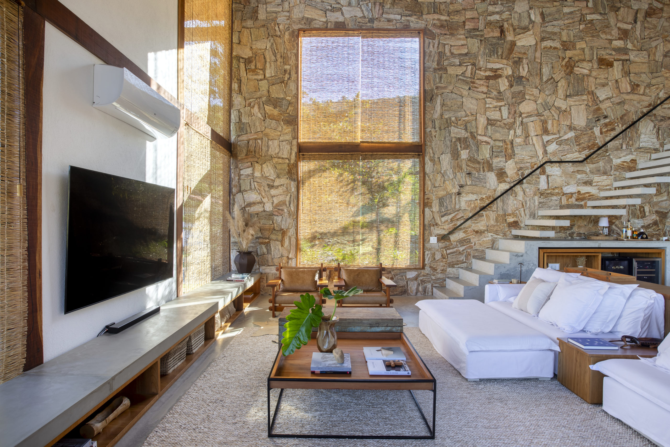 na foto uma sala de TV com escada, parede em pedras 