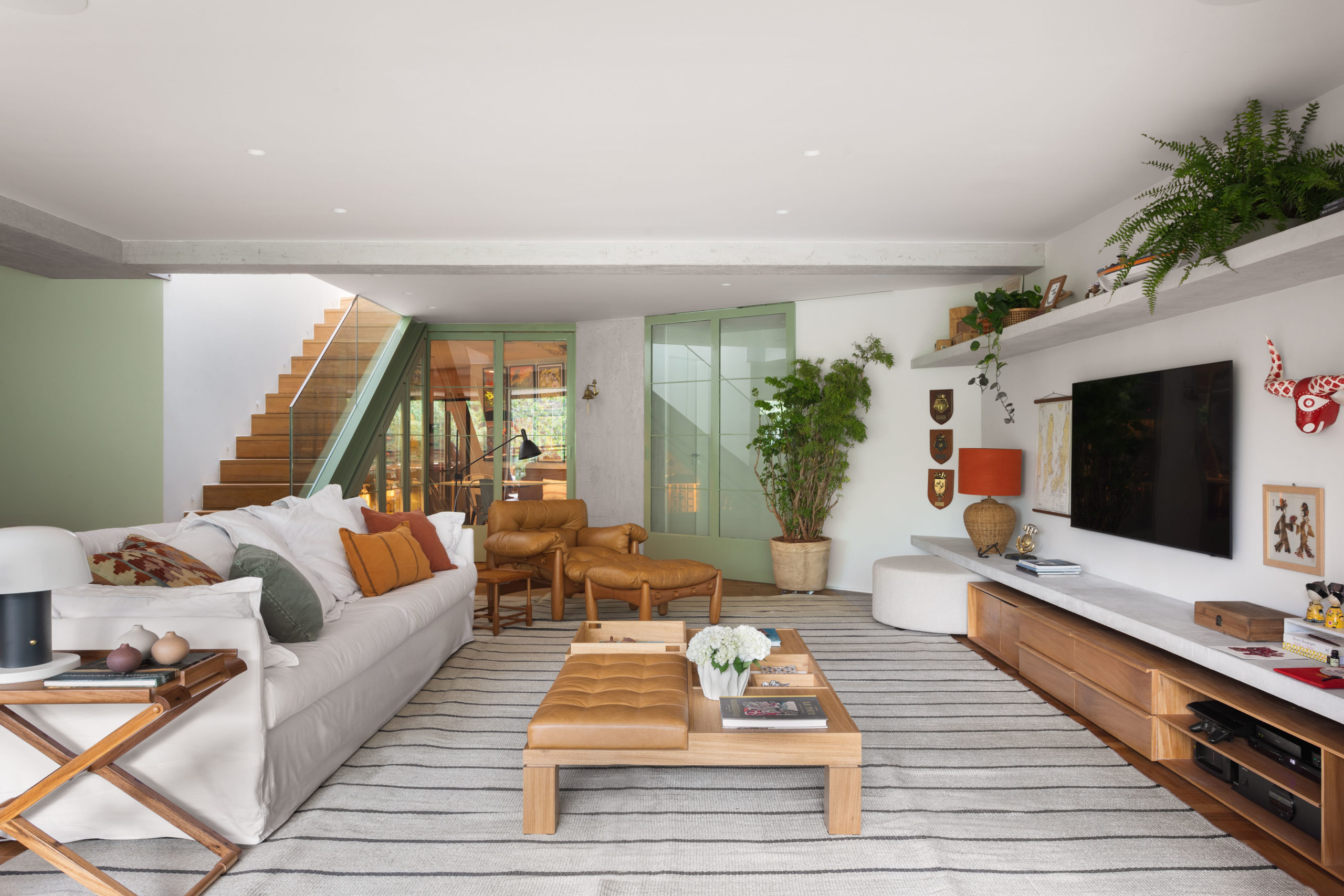 na foto uma sala com sofá, mesa de centro, tv na parede uma escada ao fundo e porta de vridro