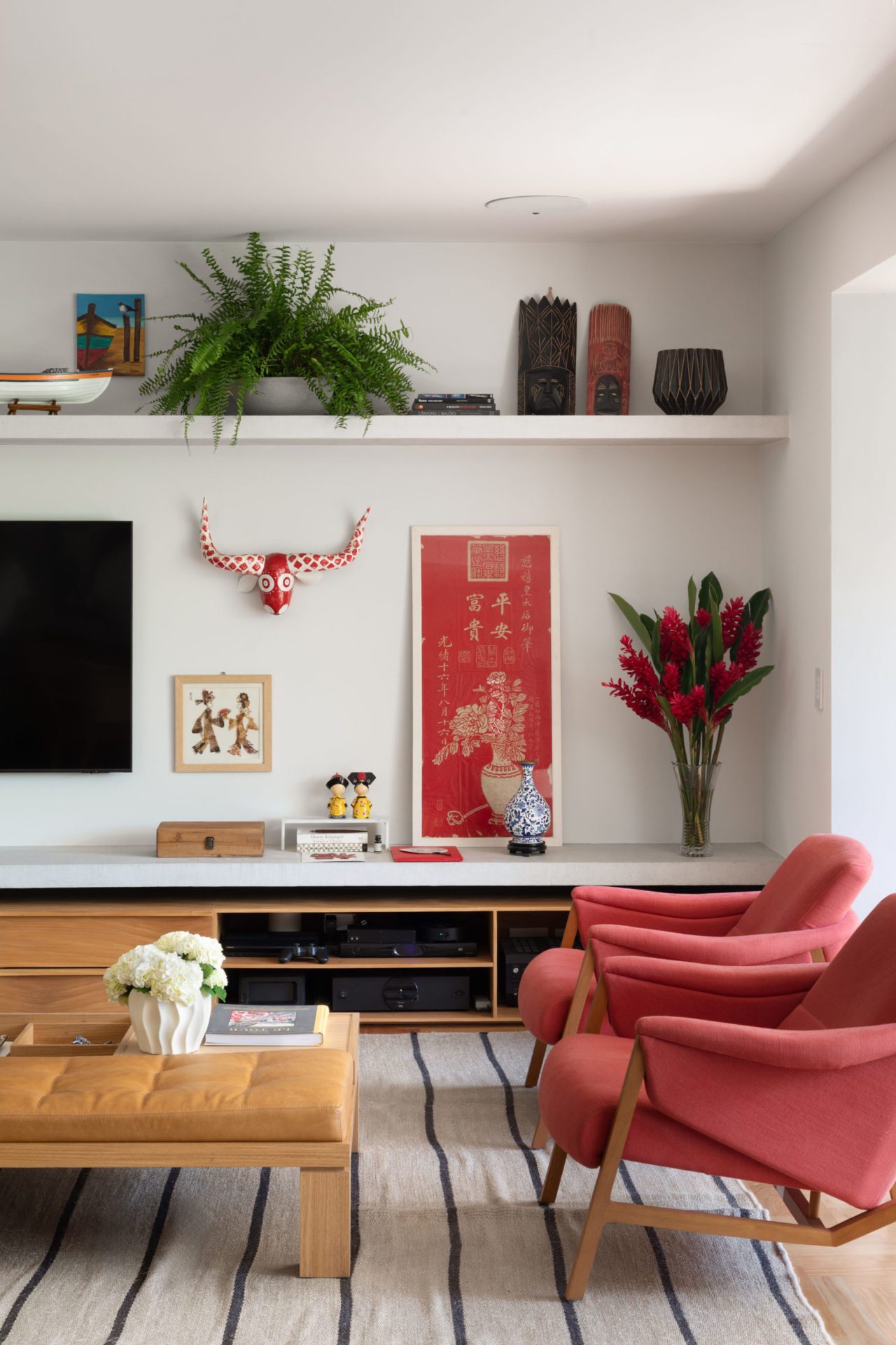 na foto sala com duas poltronas na cor cereja, rack para tv, vaso de flores vermelhas e prateleira branca