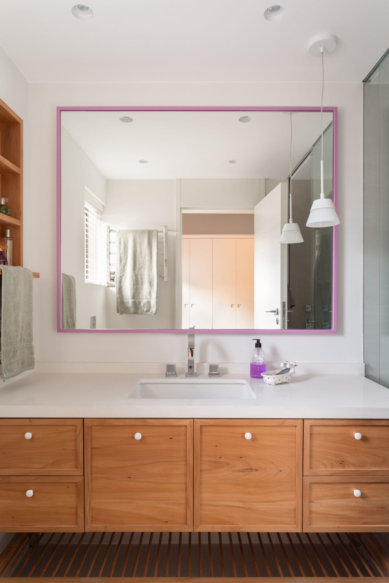na foto bancada de banheiro em madeira, espelho com moldura rosa, luminária