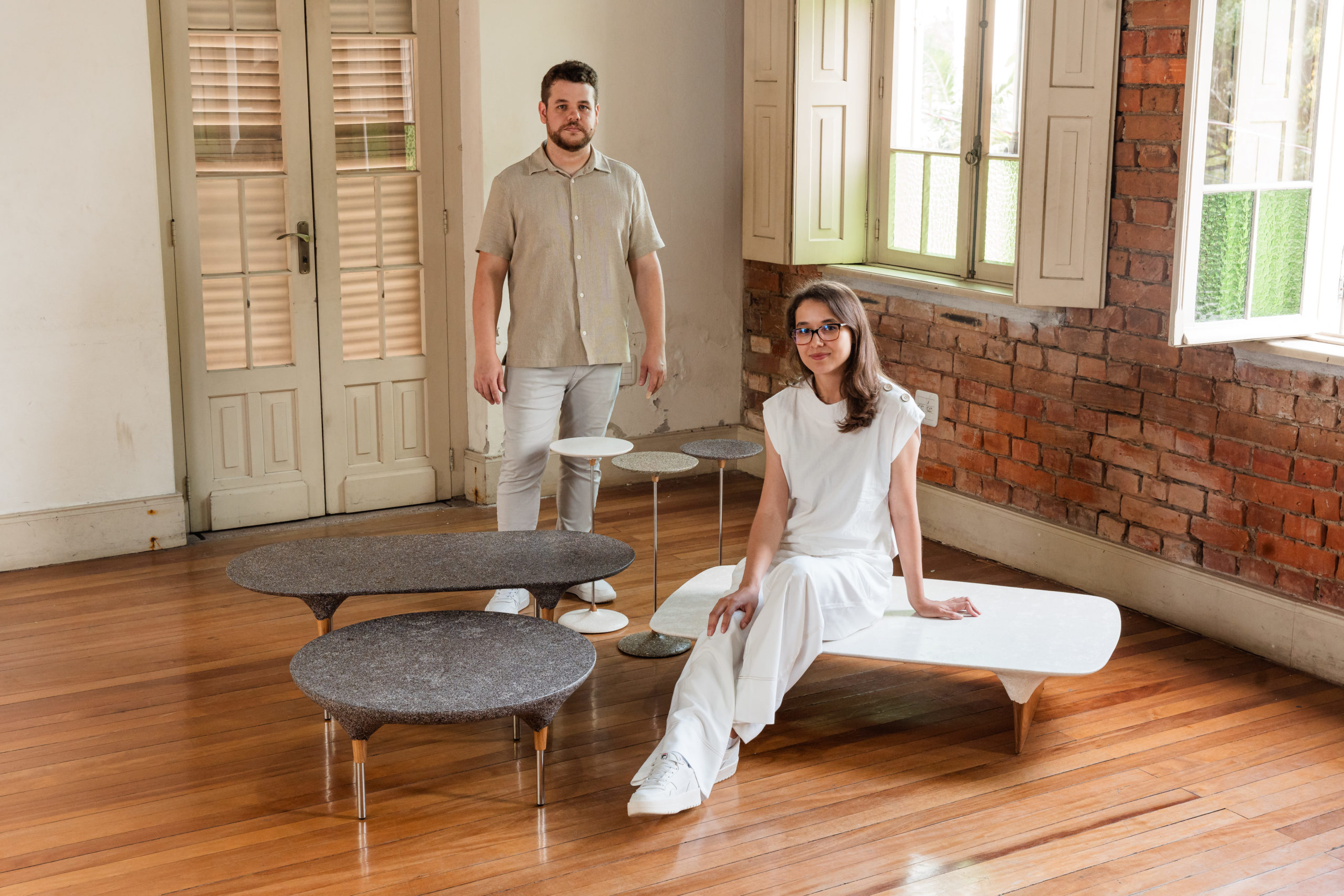 na foto uma sala com uma mulher branca de cabelos longos sentada em uma mesa baixa, um homem branco em pé e mesas de apoio