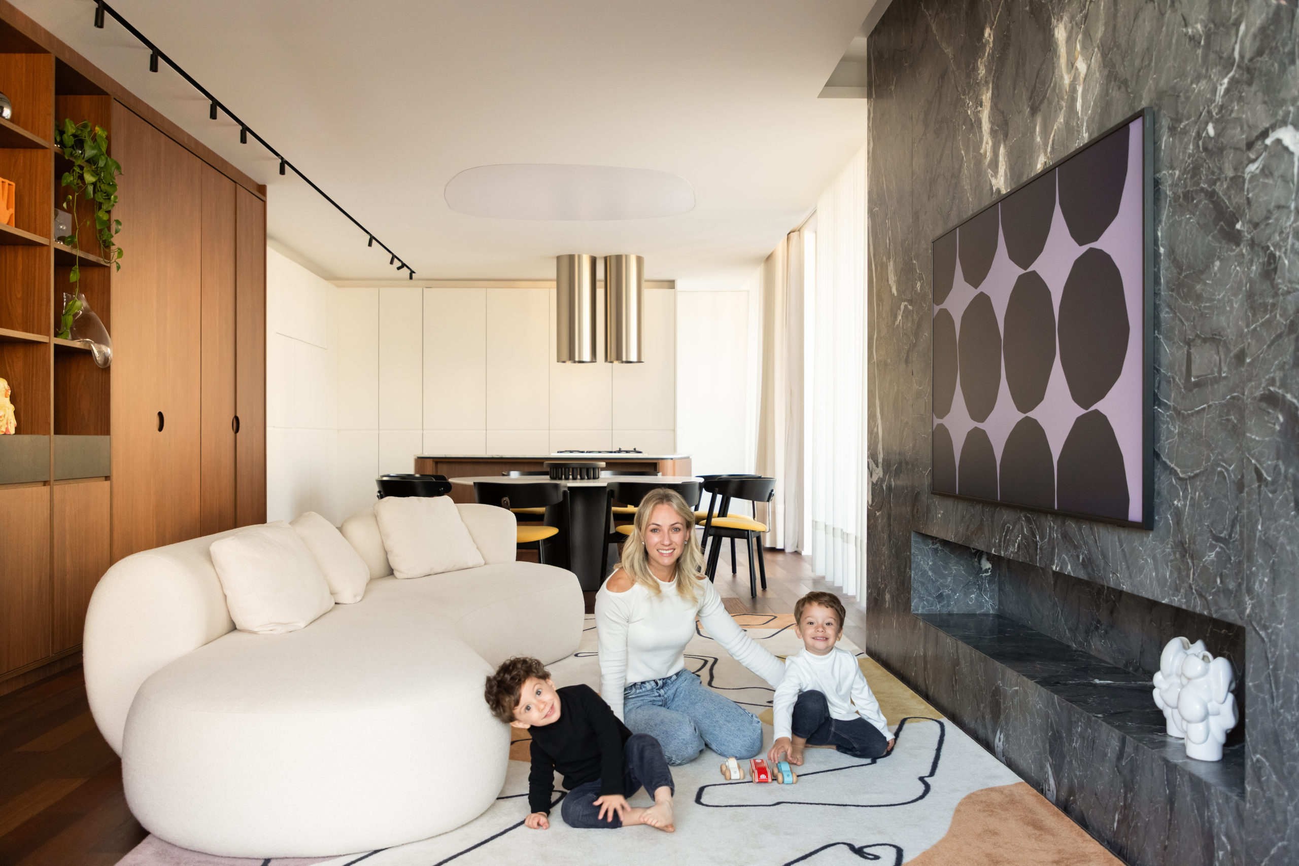 na foto uma mulher loira e branca sentada no chão da sala ao lados dos filhos gêmeos